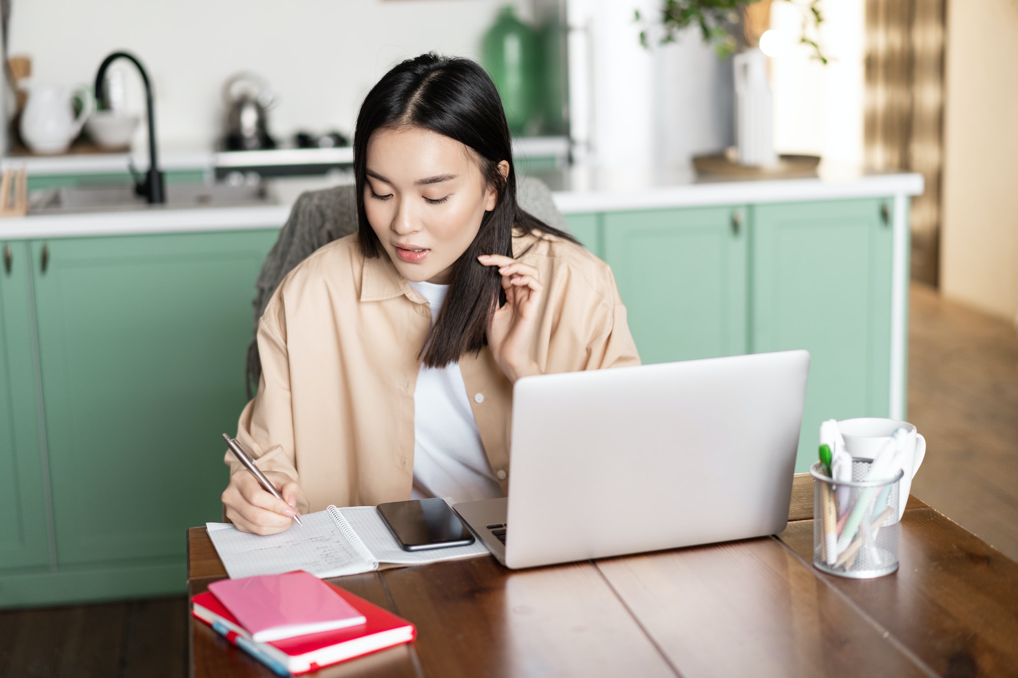 Asian girl student doing homework at home. Young woman taking notes, working on laptop and writing