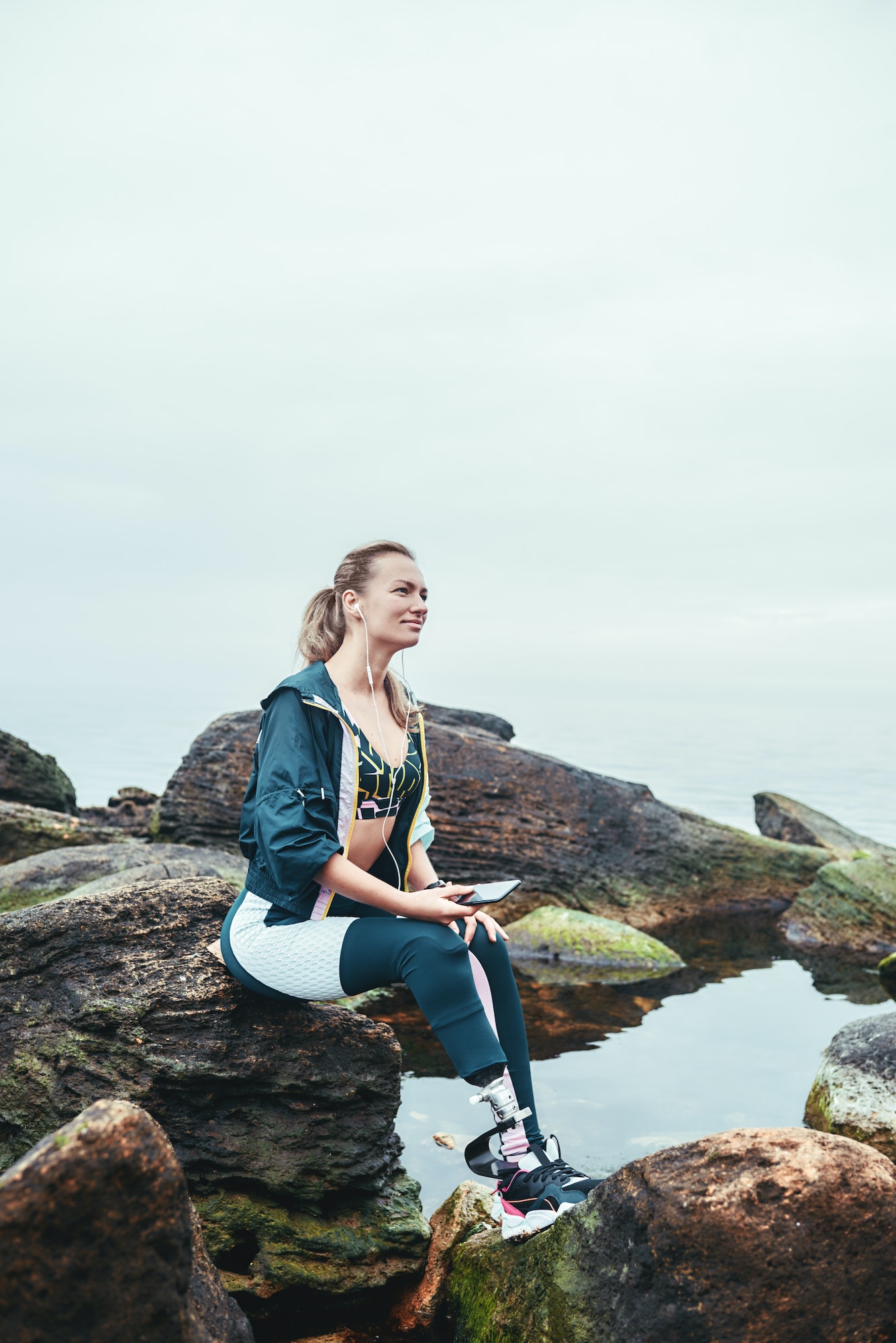 Enjoying nature and music. Happy disabled athlete woman in sportswear with prosthetic leg sitting on